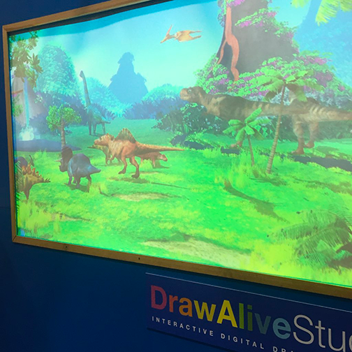 Animated dinosaur scene projected on the Draw Alive Studio screen at the IMAG